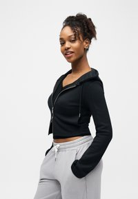 Jeune femme aux cheveux bouclés portant un sweat à capuche noir zippé et un pantalon de survêtement gris clair, debout les mains dans les poches, sur fond blanc.