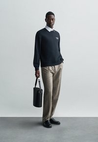 Sudadera negra con logo blanco, camisa blanca de cuello por debajo, pantalones de mezclilla beige con patrón, zapatos de cuero negros y una bolsa cilíndrica negra.