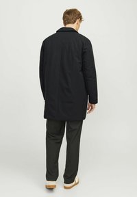 Abrigo impermeable negro con cuello y mangas largas, de corte recto. Lleva encima pantalones oscuros y zapatos de suela clara.