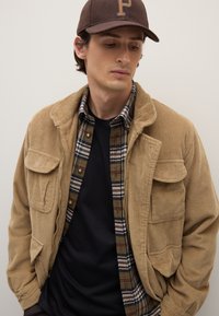 Giacca di velluto a coste color cachi con tasche sul petto, indossata sopra una camicia a quadri verde, blu scuro e bianca. T-shirt nera sotto; cappello marrone scuro.