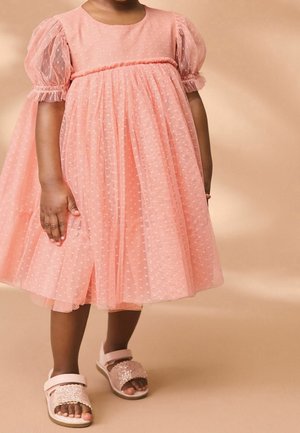 Robe en tulle rose avec de courtes manches bouffantes et une texture à pois. Associée à des sandales roses ornées d'accents scintillants.