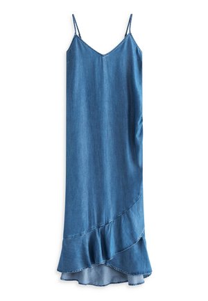 Femme portant une robe longue bleue sans manches style portefeuille et des sandales marron, debout sur une plage de sable avec des vagues de l'océan en arrière-plan.