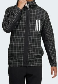 Musta takki, jossa on ruutukuosi, vetoketju edessä, huppu ja kolme valkoista raitaa rinnassa. Materiaali näyttää sileältä ja kevyeltä.