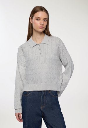 Giovane donna con capelli marroni lisci che indossa un maglione polo a costine grigio chiaro e jeans blu scuri, in piedi davanti a uno sfondo semplice.