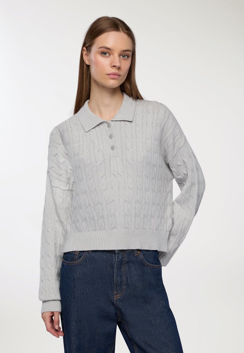 Młoda kobieta z prostymi brązowymi włosami, ubrana w jasnoszary sweter polo z splotem warkoczowym i ciemnoniebieskie dżinsy, stojąca na jednolitym tle.