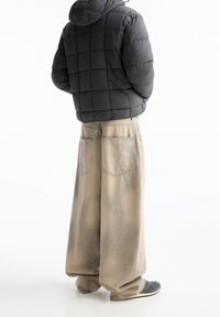 Schwarze gestepptte Daunenjacke mit Kapuze, kombiniert mit übergroßen beigen Jeanshosen mit verwaschener Textur und sichtbaren Nähten. Schwarze Schuhe.