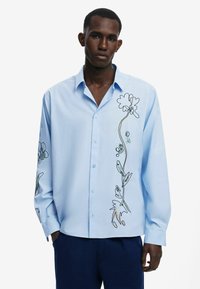 Light blue long-sleeve shirt featuring floral embroidery in black and green outlines along the left side, button-up front, and relaxed fit.