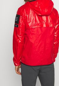 Rød vandtæt jakke med hætte, blank tekstur og et sort mærke på venstre ærme. Har elastisk kant og manchetter.