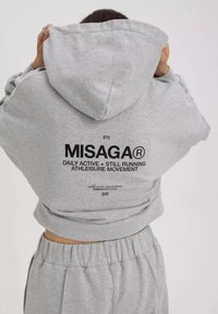 Grijze cropped hoodie met een capuchon, voorzien van zwarte bedrukte tekst op de achterkant, en bijpassende grijze broek met een elastische tailleband.