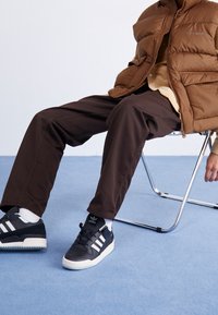 Braune Pufferjacke, beiges Hemd, dunkelbraune Hosen und schwarze Adidas-Sneaker mit weißen Streifen. Glatte Texturen; minimalistisches Design.