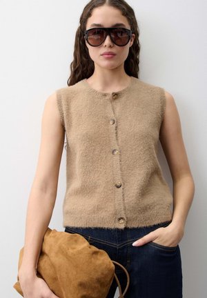 Donna con grandi occhiali da sole neri, top beige senza maniche peloso con bottoni, jeans scuri, che tiene una borsa marrone in suede in una mano.