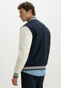 Giacca bomber di un blu scuro con maniche bianche, polsini a coste e colletto a righe. Presenta un materiale testurizzato con un design informale e sportivo.
