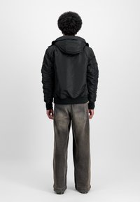 Veste isolante noire avec capuche, présentant des poignets côtelés et une coupe décontractée. Portée avec un jean gris délavé à jambes larges.