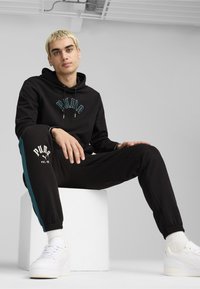 Felpa nera con logo ricamato "PUMA", abbinata a pantaloni jogger neri con pannelli laterali color teal. Indossato con sneakers bianche, seduto su un blocco bianco.