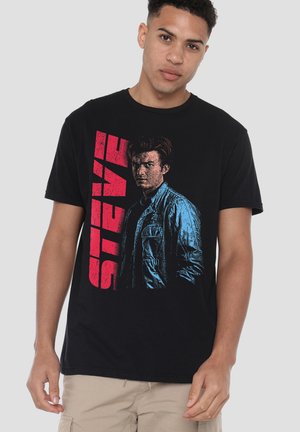 Schwarzes Baumwoll-T-Shirt mit einer großen Retro-Grafik einer Person in einer blauen Jacke und dem Wort "STEVE" in fetten roten Buchstaben.