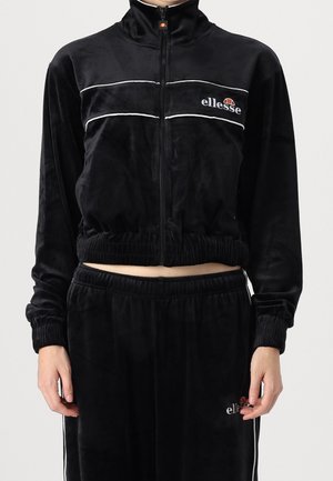 Personne portant un survêtement en velours noir ellesse avec une veste zippée et un pantalon à ceinture élastique, avec un liseré blanc et un logo sur la poitrine et la cuisse.
