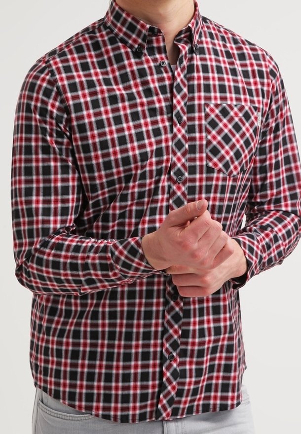 Mies, joka on pukeutunut punaisiin, mustiin ja valkoisiin ruudullisiin pitkähihaisiin paitoihin, puristaa yhtä kättään toisen edessä.