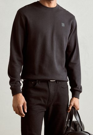 Homme portant un sweat-shirt noir à manches longues et un pantalon noir, tenant un sac en cuir noir dans sa main droite sur un fond uni.