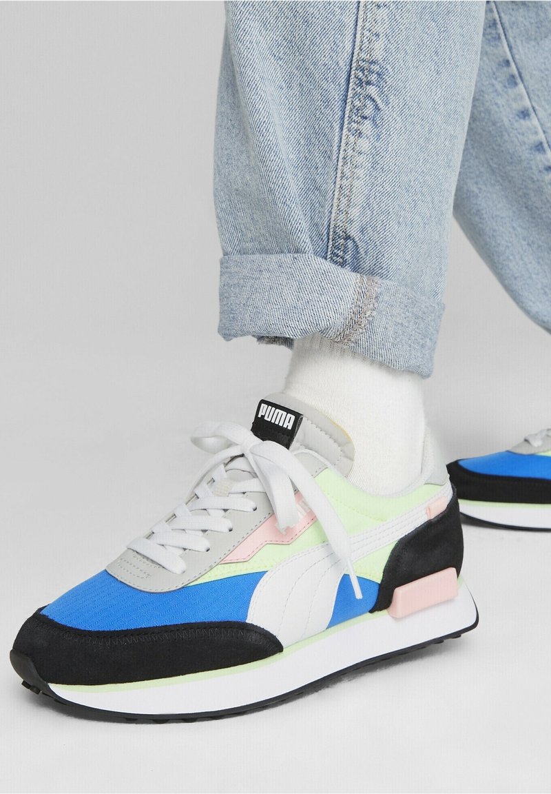 Bunt gefärbter Sneaker mit blauen, grünen, schwarzen und pinken Paneelen, einem weißen Logo und einer strukturierten Sohle, kombiniert mit hochgerollten Jeans.