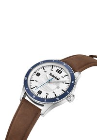 Reloj de acero inoxidable con un dial blanco, bisel azul, correa de ante marrón y marcadores de hora prominentes. Presenta un logo y corona plateados.