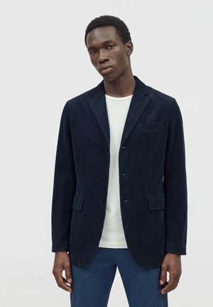 Jeune homme portant un blazer en velours côtelé bleu marine sur une chemise blanche et un pantalon bleu, debout devant un fond clair uni.