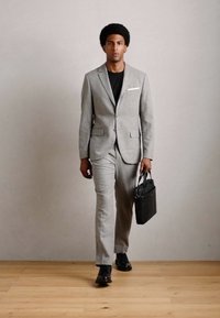 Mann som går innendørs iført en lys grå dress og svart skjorte, holder en svart dokumentmappe, på tregulv mot en ensfarget vegg.