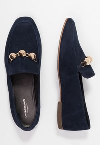 Mocasines de ante azul marino con puntera cuadrada, que presentan acentos circulares en tono dorado y un tacón bajo marrón. Textura suave con detalles de costura mínimos.