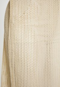 Tessuto lavorato a maglia beige con una trama aperta, caratterizzato da una texture leggera e ariosa. Il design si drappeggia dolcemente con una forma rilassata.