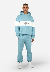 Svetlomodrá mikina s bielym horizontálnym pruhom, na ktorej je nápis "Pegador". Mäkká látka, predná vrecko, v kombinácii s odpovedajúcimi jogger nohavicami. Biely športový obuv.