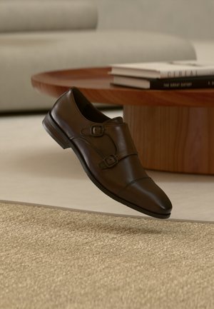 Chaussure habillée en cuir marron foncé à double boucle flottant au-dessus d'un tapis beige texturé dans un salon moderne.