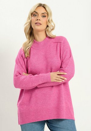Femme aux cheveux blonds portant un pull rose et un jean bleu, debout les bras croisés devant un fond clair uni.