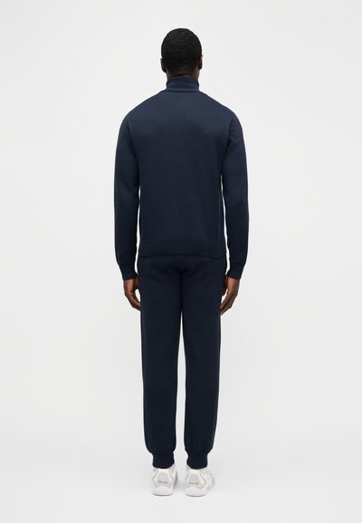 Vyras stovintis nugara, dėvintis tamsiai mėlyną golfą ir derančias jogger stiliaus kelnes bei baltus sportbačius.