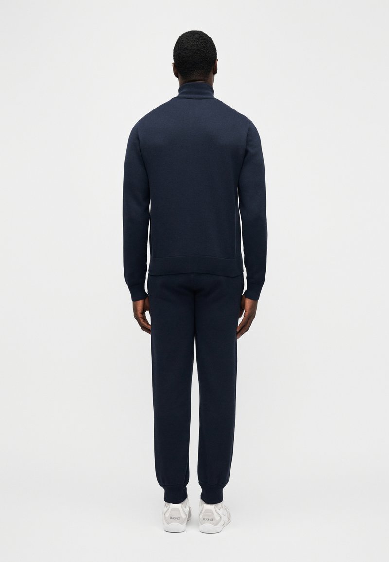 Homme debout, vu de dos, portant un pull à col roulé bleu marine et un pantalon de jogging assorti avec des baskets blanches.