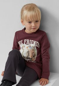 Młoda blondynka siedzi, ubrana w bordo bluzę z napisem "GIRL FRIENDS" i ilustracjami koni, w połączeniu z ciemnymi spodniami i czarnymi butami.