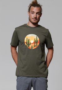 Olijfgroen katoenen t-shirt met een cirkelvormige afbeelding van cactussen en een zonsondergang in oranje en gele tinten. Ronde hals en korte mouwen.