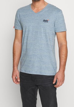 Uomo con t-shirt a maniche corte azzurra con scollo a V e piccolo logo sul petto, pantaloni neri, in piedi davanti a uno sfondo neutro.