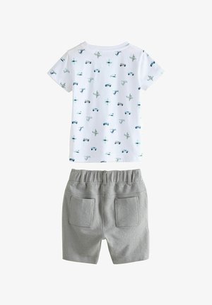 Īsām piedurknēm baltā T-kreklā ar zilu transportlīdzekļu un zvaigžņu rakstu, savienots ar pelēkām biksēm, kuru josta ir elastīga un kurām ir divas aizmugures kabatas.