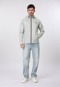 Άνδρας που φοράει ανοιχτό γκρι φούτερ με φερμουάρ, ανοιχτό μπλε τζιν και λευκά αθλητικά παπούτσια, στέκεται με το ένα χέρι στην τσέπη μπροστά σε απλό φόντο.