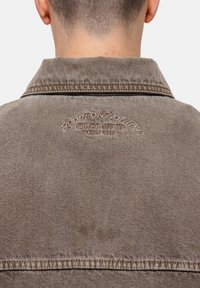 Bruine denimjas met een kraag, met geborduurde tekst op de rug. Zichtbare textuur en stiksels langs de randen. Casual ontwerp.