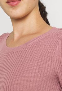 Pull en maille côtelée rose clair avec encolure ronde et texture subtile ; tissu doux présentant de fines lignes verticales.