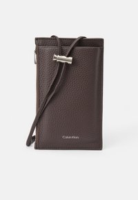 Brązowy skórzany portfel na karty z zamkiem błyskawicznym, regulowanym paskiem i srebrnymi detalami. Wytłoczone logo Calvin Klein z przodu.