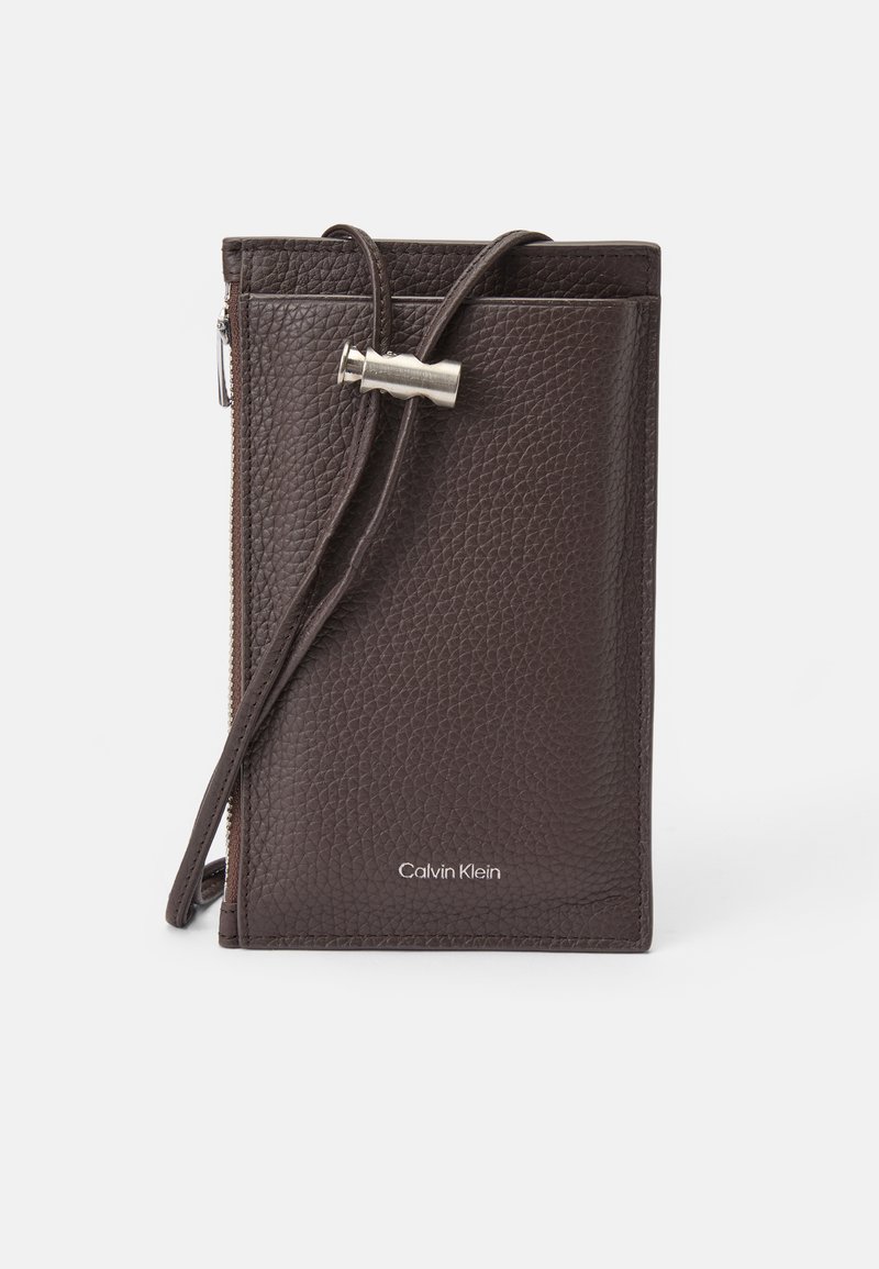 Brązowy skórzany portfel na karty z zamkiem błyskawicznym, regulowanym paskiem i srebrnymi detalami. Wytłoczone logo Calvin Klein z przodu.
