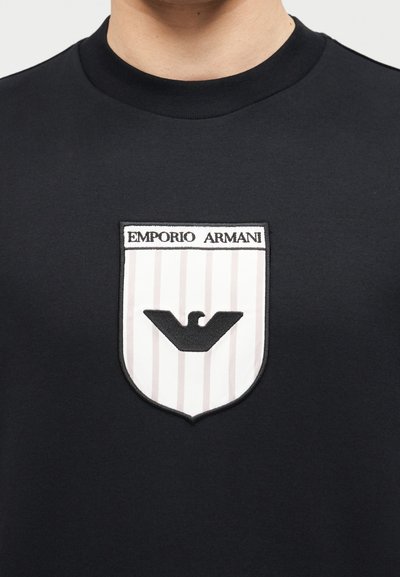 Μαύρο πουκάμισο με ένα λευκό και ανοιχτό γκρι ριγέ έμβλημα σε σχήμα ασπίδας που απεικονίζει έναν μαύρο αετό και την επιγραφή «EMPORIO ARMANI» στο στήθος.