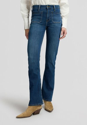 Vrouw met donkerblauwe bootcut jeans, beige suède puntlaarzen en een wit shirt met lange mouwen, staand op een grijze achtergrond.