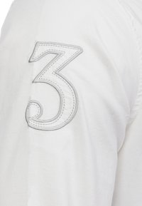 Bílý rukáv košile s velkým šedým vyšívaným číslem "3" s zatočeným akcentem na hladké textuře látky.