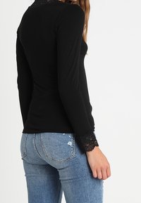 Schwarzes langärmeliges Oberteil mit Spitzenakzenten am Kragen und an den Manschetten, kombiniert mit blauen, ausgewaschenen Jeans. Figurbetontes Design, glatte Textur.