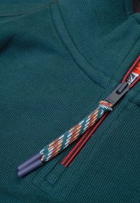 Groene textuurstof met een rode rits. Heeft een decoratieve trekkoord met diagonale patronen in oranje, wit en groen.