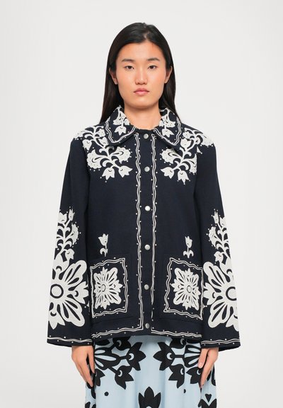 Veste marine avec broderie florale blanche, présentant un grand col, des boutons à l'avant et des manches longues. Ourlet effilé avec des accents détaillés.