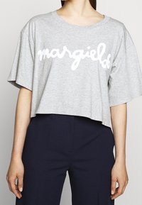 Modell iklädd en grå kortärmad t-shirt med texten "margielof" i vitt, tillsammans med mörkblå byxor, mot en neutral bakgrund.