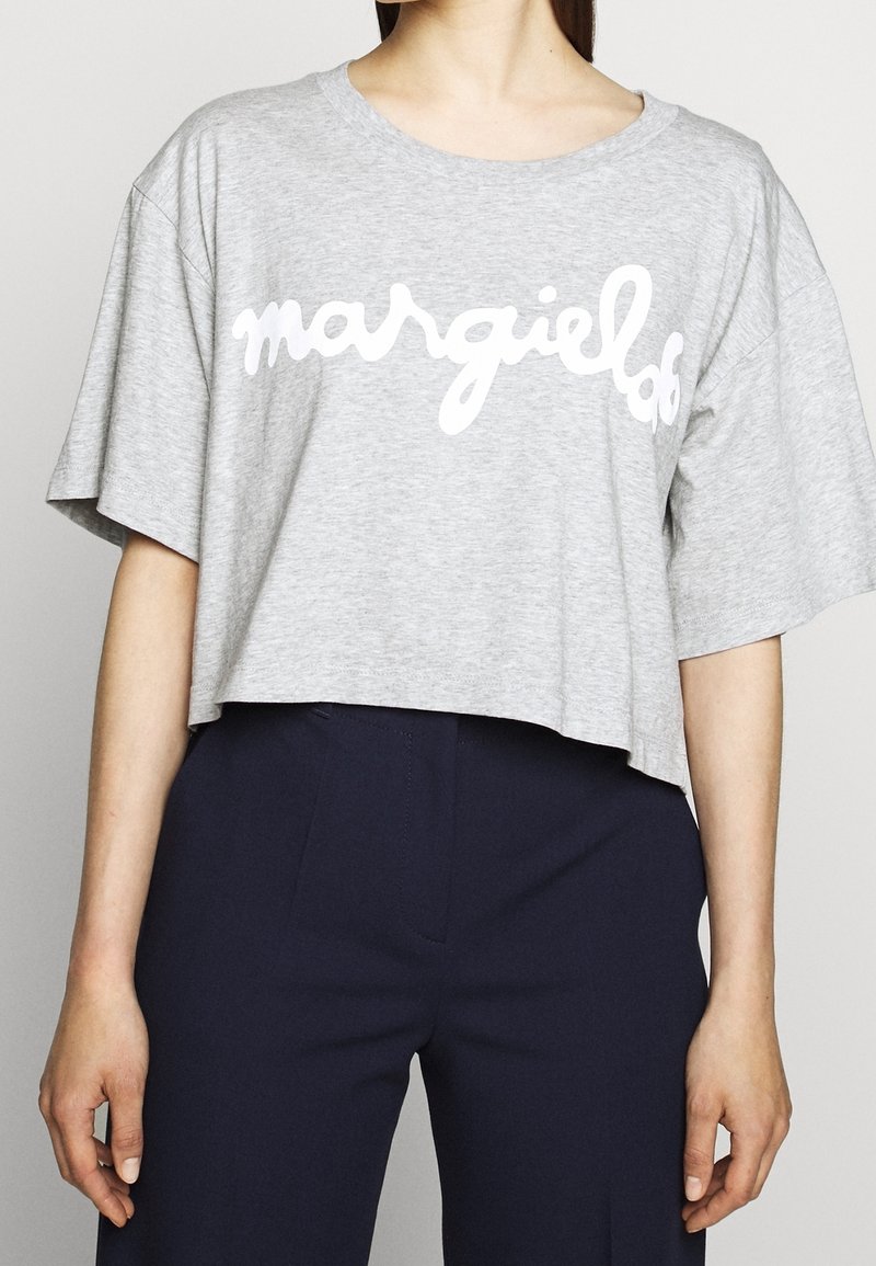 Modell iklädd en grå kortärmad t-shirt med texten "margielof" i vitt, tillsammans med mörkblå byxor, mot en neutral bakgrund.